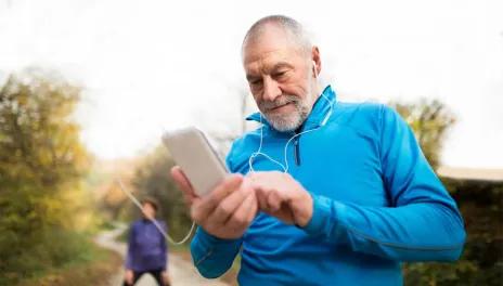 An older man standing on a running path looking at his mobile device. He's wearing athletic clothing and earbuds.