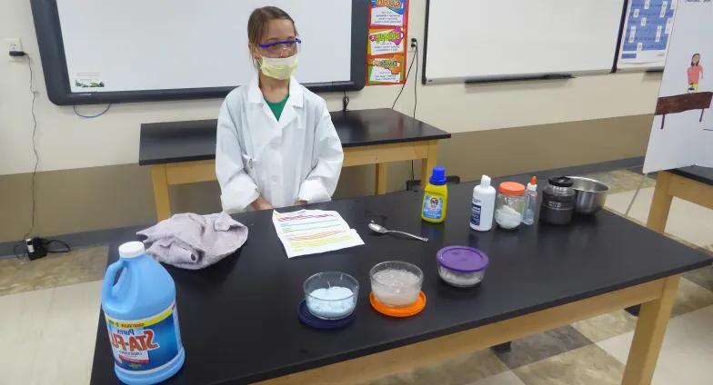 girl with mask and science lab coat on standing behind table with experiment ingredients