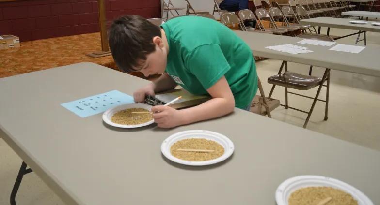 boy writing on sheet of paper with paper plates of seeds in front of him on table