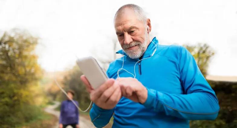 An older man standing on a running path looking at his mobile device. He's wearing athletic clothing and earbuds.